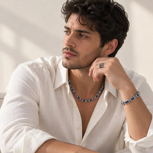 A man wearing a silver-toned chain necklace and matching bracelet with rectangular blue gemstones, styled with an open white shirt against a softly lit neutral background.