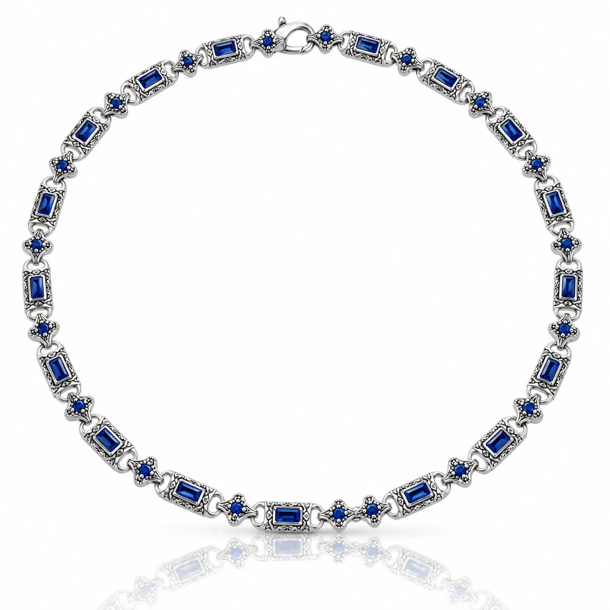 A silver-toned tennis necklace featuring rectangular and round-cut deep blue gemstones, displayed on a clean white background with a soft reflection below.