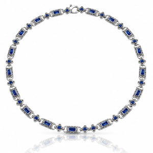 A silver-toned tennis necklace featuring rectangular and round-cut deep blue gemstones, displayed on a clean white background with a soft reflection below.