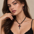 A close-up of a woman wearing a gothic-style black cord necklace with a silver-toned cross pendant featuring a red rose, along with matching drop earrings.