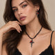 A close-up of a woman wearing a gothic-style black cord necklace with a silver-toned cross pendant featuring a red rose, along with matching drop earrings.