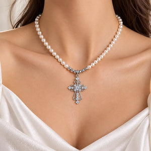 A close-up of a woman's neck and upper chest wearing a pearl necklace with a silver-toned cross pendant adorned with clear stones, paired with a white satin top.