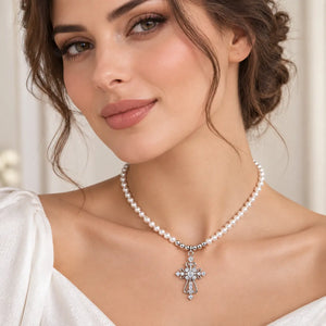 A close-up of a woman wearing a delicate white pearl necklace with a silver-toned cross pendant adorned with sparkling clear stones, set against a soft, light beige background.