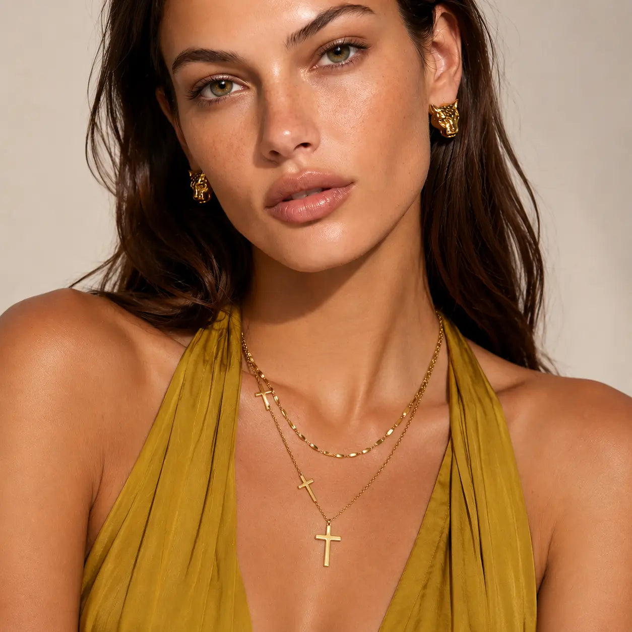 A close-up of a woman wearing gold cross pendants on layered necklaces and matching gold hoop earrings, paired with a mustard yellow halter top against a neutral beige background.