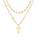 A gold layered necklace set on a white background, featuring a short chain with a small cross, a longer chain with a larger cross pendant, and a beaded chain in the back.