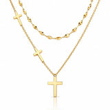 A gold layered necklace set on a white background, featuring a short chain with a small cross, a longer chain with a larger cross pendant, and a beaded chain in the back.