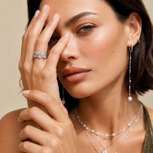A close-up lifestyle shot of a model wearing a silver patterned ring, a long silver dangle earring with a pearl, and layered silver pearl necklaces against a soft beige background.