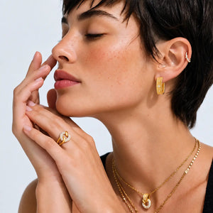 A close-up lifestyle shot of a woman with short dark hair wearing gold jewelry, including a gold hoop earring, layered gold necklaces, and a gold ring with a clear gemstone accent.