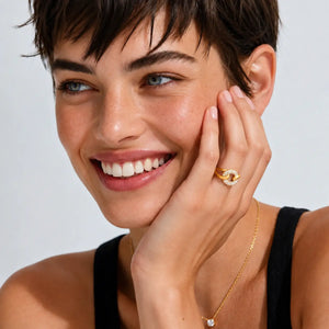 A close-up lifestyle shot of a smiling woman with short brown hair, wearing a gold ring with a white stone and a delicate gold chain necklace. She is touching her cheek, showcasing the jewelry against a clean, light gray background.