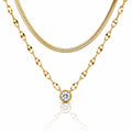 A gold necklace set featuring a double-layered chain design: a thinner snake chain and a thicker oval-link chain with a round bezel-set clear gemstone pendant, displayed on a white background with a soft reflection.