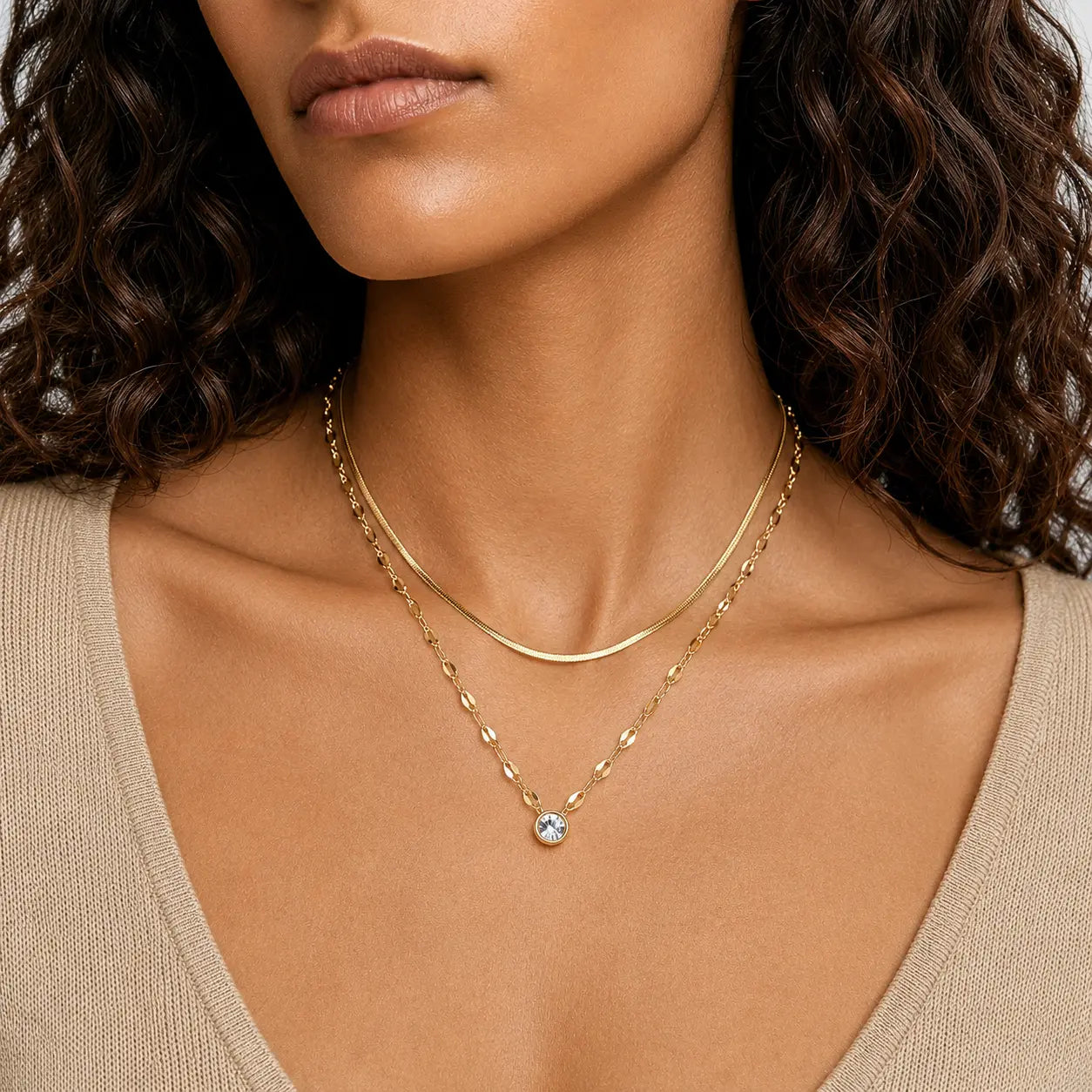 A close-up of a model wearing a delicate gold double-layer necklace with a small clear crystal pendant, paired with a beige top.