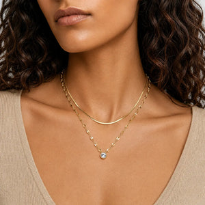 A close-up of a model wearing a delicate gold double-layer necklace with a small clear crystal pendant, paired with a beige top.