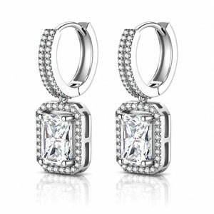 A pair of silver-toned huggie hoop earrings with rectangular emerald-cut clear gemstones surrounded by small pavé-set stones, shown on a clean white background with a soft reflection below.