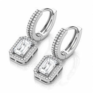 A pair of silver-toned huggie hoop earrings with rectangular emerald-cut clear stones and halo accents, displayed on a clean white background with a soft reflection.