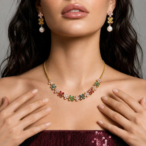 A close-up lifestyle shot of a model wearing a gold floral necklace and matching drop earrings. The necklace and earrings feature gold flower-shaped settings adorned with small multicolored gemstones (pink, green, blue, red, and white) and a single white pearl hanging below each flower.