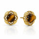 A pair of gold stud earrings featuring round brown tiger's eye centers surrounded by a halo of small clear stones and a scalloped gold setting, shown on a white background with a soft reflection.