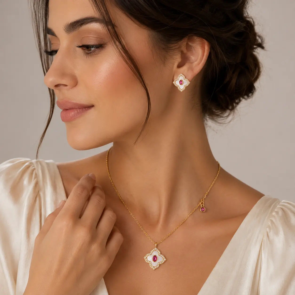 A woman wearing a gold necklace and matching stud earring with white enamel and a small red gemstone, shown in a close-up portrait against a soft neutral background.