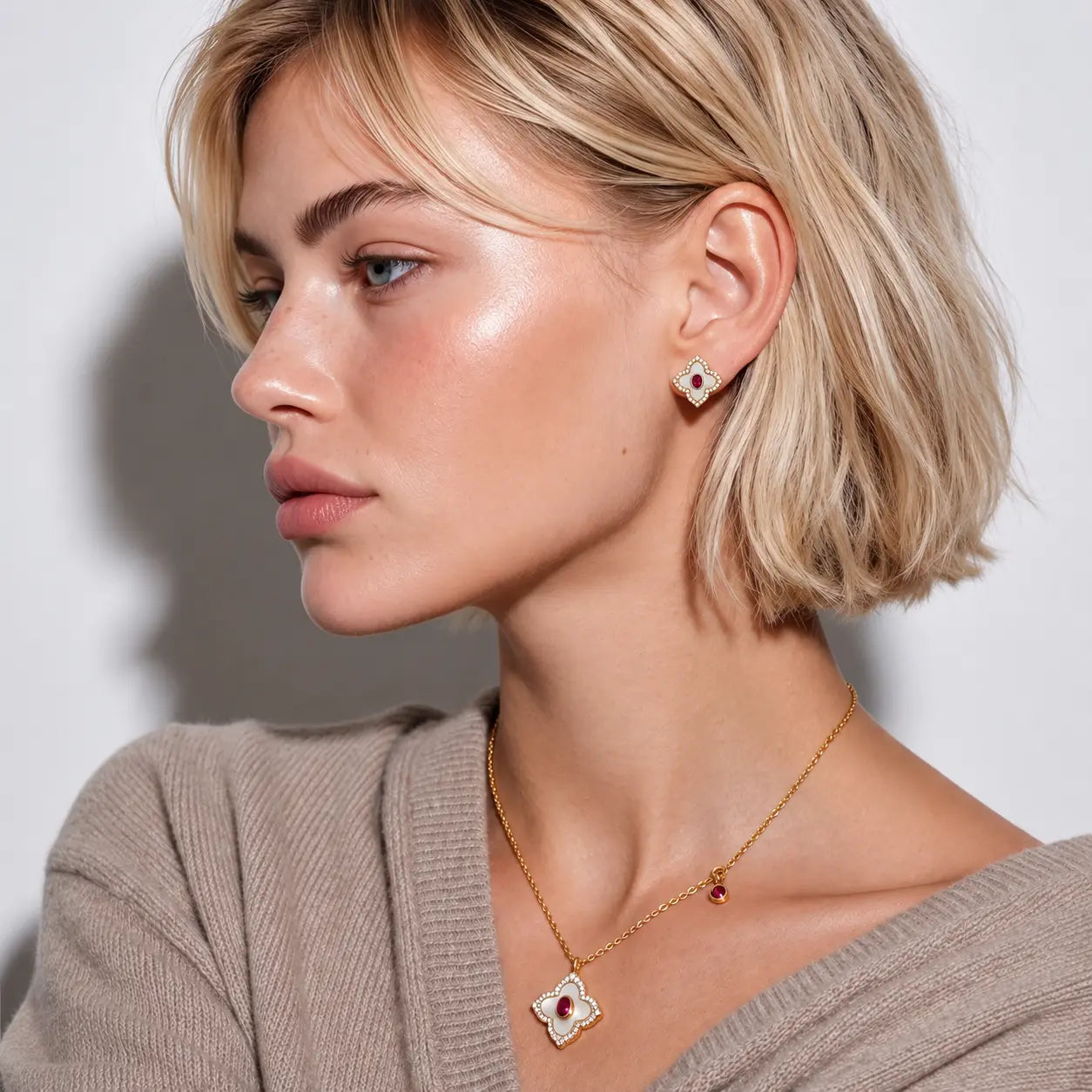 A close-up side profile of a blonde woman wearing a delicate gold necklace with a red and white clover pendant and matching stud earrings, paired with a beige knit sweater against a neutral light gray background.