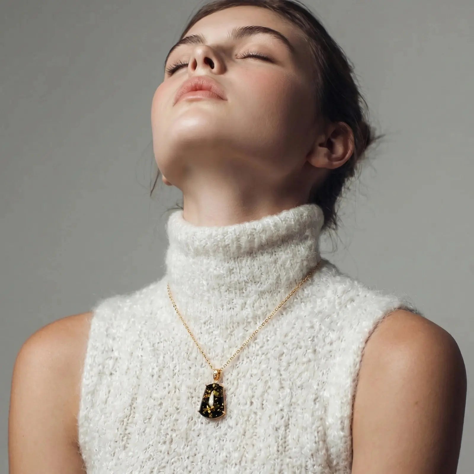 Woman wearing a white turtleneck sweater and gold necklace with a dark pendant against a gray background