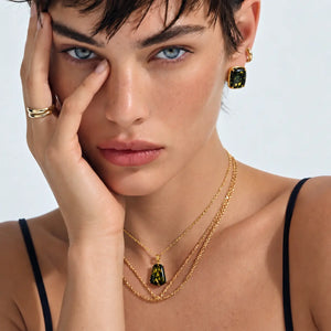 A close-up lifestyle shot of a model wearing a gold layered necklace set with green rectangular gemstone pendants and matching drop earrings.