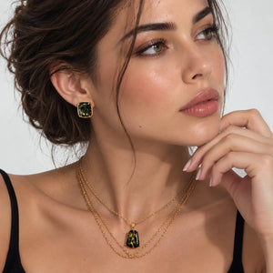 A close-up lifestyle shot of a woman wearing a matching gold jewelry set, featuring a gold pendant necklace with a green faceted stone and a matching gold stud earring.
