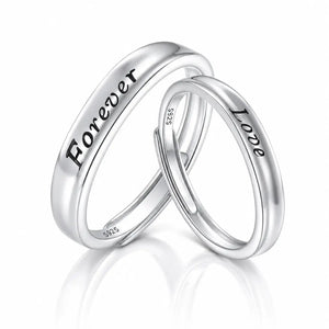 Two polished silver wedding bands interlocking on a reflective white surface. One band is engraved with the word 'Forever' and the other with 'Love'.
