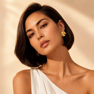 A close-up portrait of a woman with a short dark bob hairstyle wearing a textured gold hoop earring, set against a warm beige background with soft light shadows.