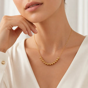 A close-up of a woman wearing a delicate gold necklace with a row of small, polished gold beads at the center, styled with a white satin blouse.