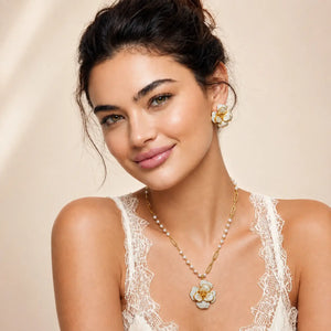 A woman wearing a gold and white flower pendant necklace with a matching flower stud earring, styled with a cream lace camisole against a soft beige background.