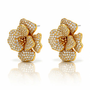 A pair of gold flower-shaped stud earrings paved with small clear stones, shown on a white background with a soft reflection below.