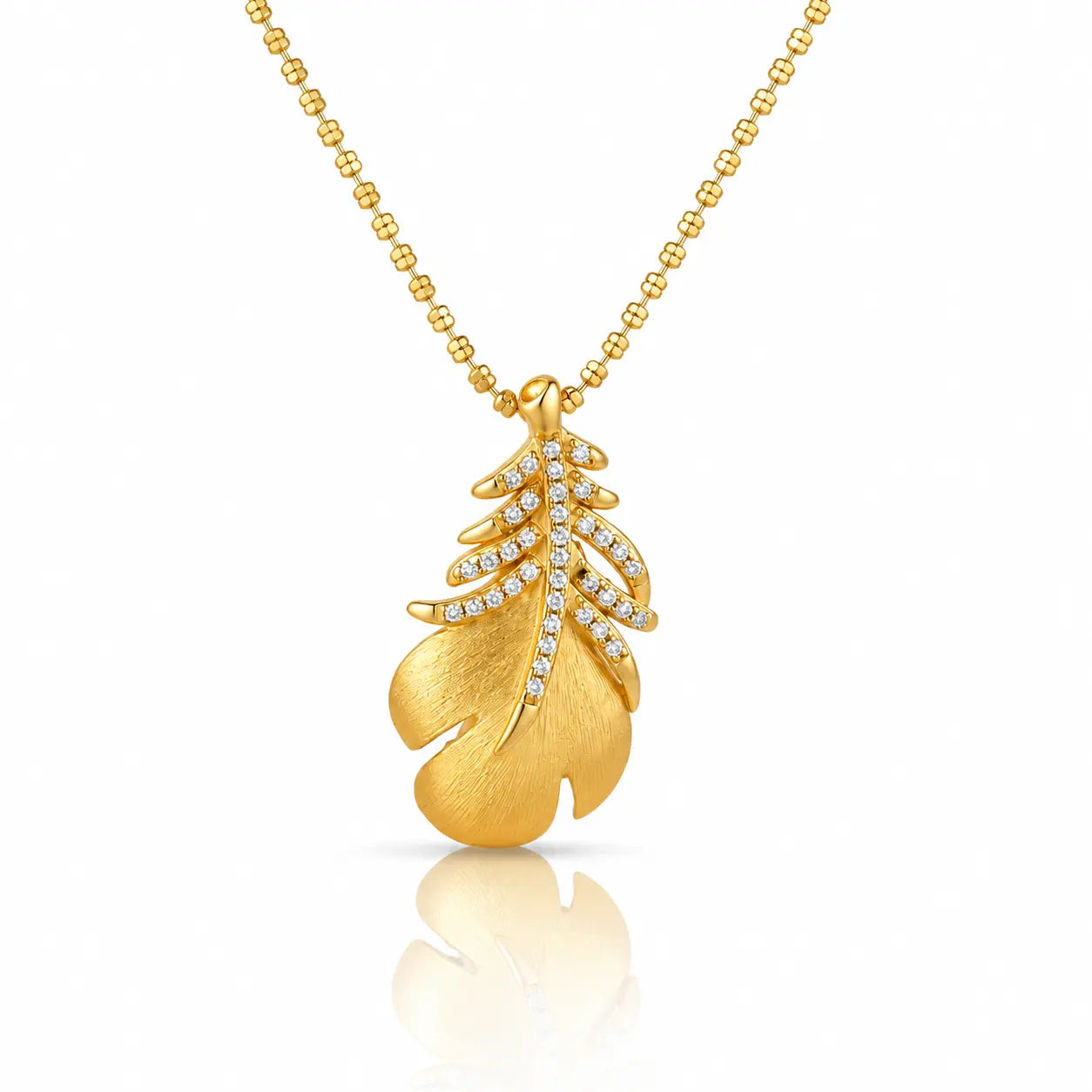 A gold pendant necklace featuring a textured leaf design adorned with small clear stones, suspended from a gold ball chain on a white background with a soft reflection below.