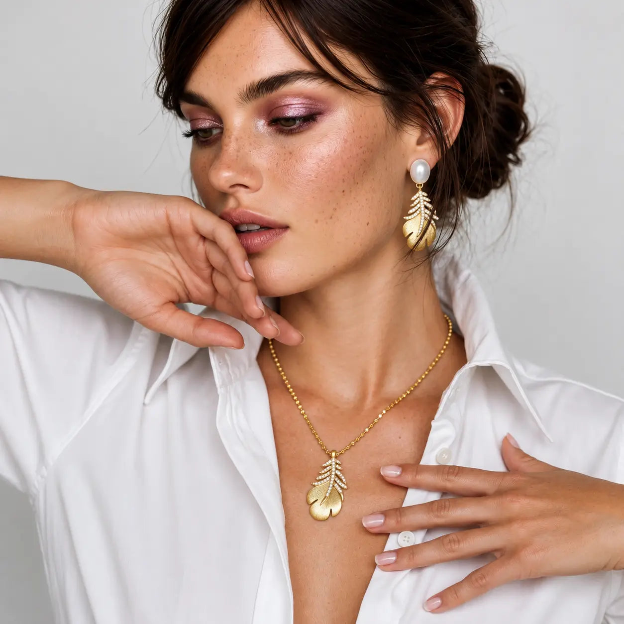 A woman wearing a gold leaf pendant necklace and matching gold leaf drop earrings with white pearl accents, styled with a white button-down shirt against a light gray background.