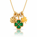 A gold St. Patrick's Day necklace featuring three shamrock charms: one large green four-leaf clover with emerald-cut stones, one solid gold shamrock, and one small gold shamrock with clear stones, on a gold ball chain.