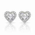 A pair of heart-shaped stud earrings featuring large clear heart-cut center stones surrounded by a halo of smaller clear stones, set in a silver-toned metal on a white background with a soft reflection below.