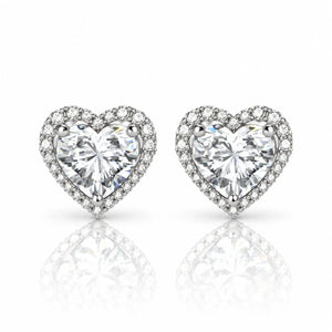 A pair of heart-shaped stud earrings featuring large clear heart-cut center stones surrounded by a halo of smaller clear stones, set in a silver-toned metal on a white background with a soft reflection below.