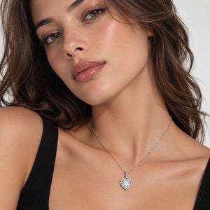 A close-up of a woman wearing a delicate silver chain necklace with a heart-shaped pendant set with clear stones, styled with a black ribbed tank top against a light gray background.