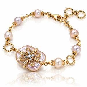 A gold chain bracelet featuring baroque-style pinkish pearl beads and a large central iridescent pearl pendant with a gold floral filigree setting and small clear stones, shown on a white reflective background.
