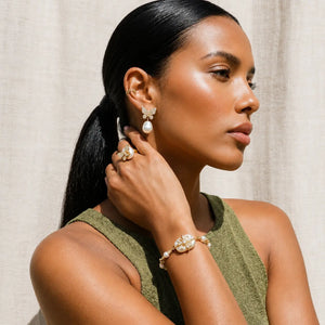 A woman with dark hair in a ponytail models a matching jewelry set featuring a pearl flower bracelet, matching pearl flower earrings, and a pearl ring, set against a neutral beige curtain background.