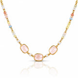 A gold-tone beaded necklace featuring three central light pink rectangular gemstones, flanked by small white pearls and multicolored beads in pastel shades of yellow, blue, and peach, set against a clean white background with a soft reflection below.