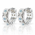 A pair of silver huggie hoop earrings decorated with white mother-of-pearl flower petals, clear rhinestones, and light blue oval center stones, displayed on a reflective white surface.