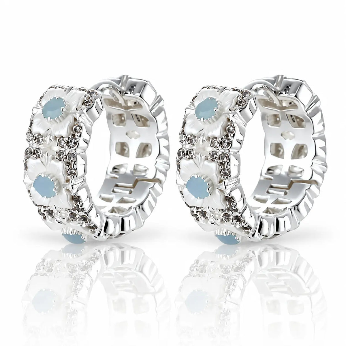 A pair of silver huggie hoop earrings decorated with white mother-of-pearl flower petals, clear rhinestones, and light blue oval center stones, displayed on a reflective white surface.
