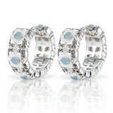 A pair of silver huggie hoop earrings decorated with white mother-of-pearl flower petals, clear rhinestones, and light blue oval center stones, displayed on a reflective white surface.