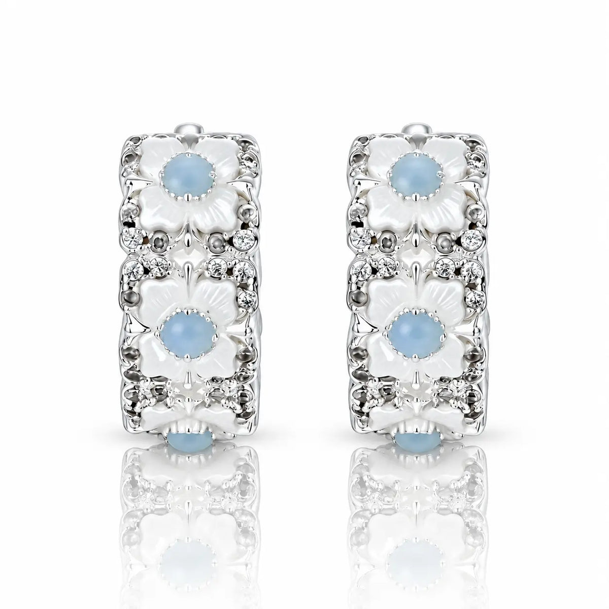 A pair of silver huggie hoop earrings decorated with three stacked white floral motifs, each featuring a light blue cabochon stone and surrounded by small clear crystals, set against a white background with a soft reflection below.