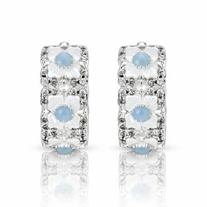 A pair of silver huggie hoop earrings decorated with three stacked white floral motifs, each featuring a light blue cabochon stone and surrounded by small clear crystals, set against a white background with a soft reflection below.