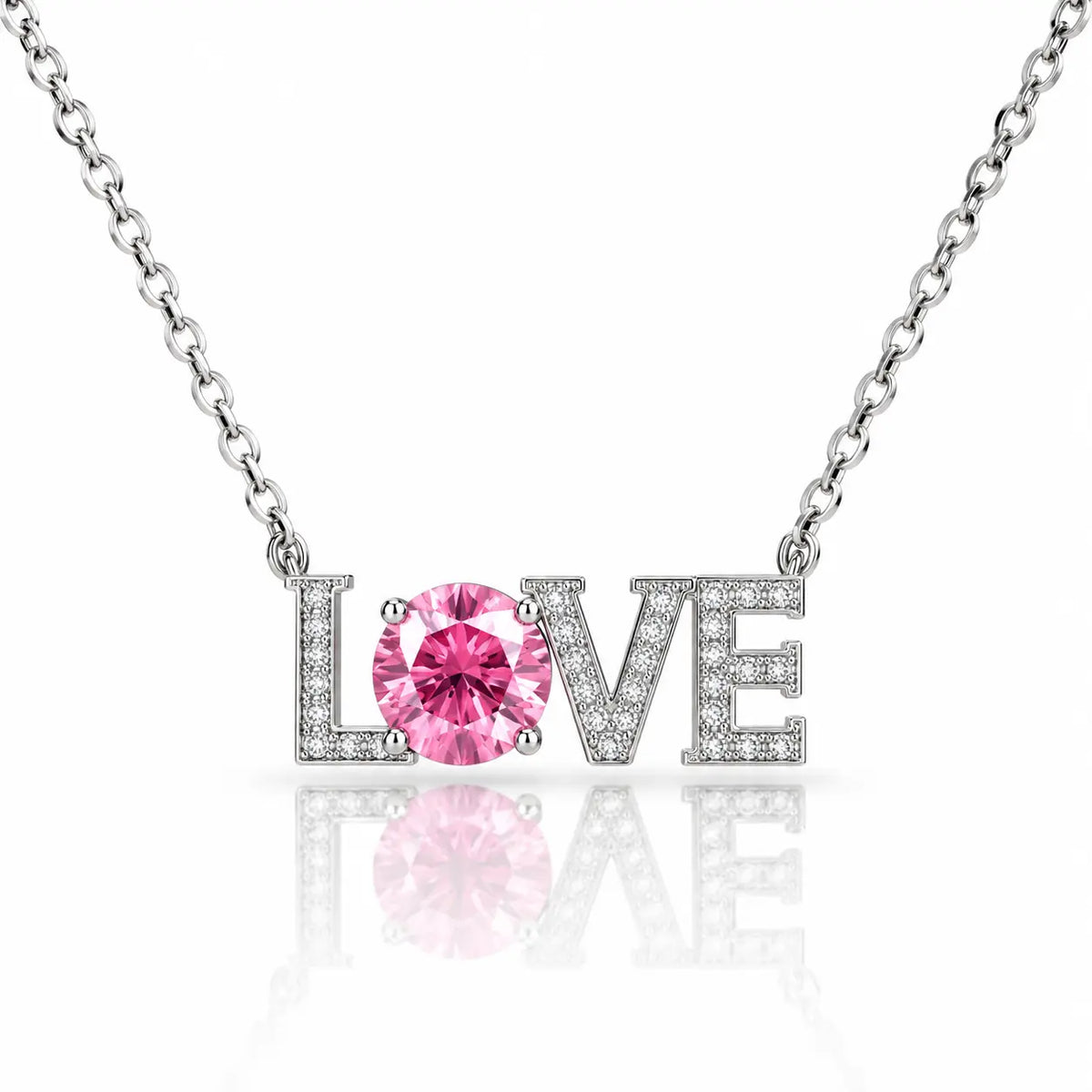 A silver-tone necklace with a pendant spelling LOVE, where the O is a round pink gemstone and the other letters are set with small clear stones, on a white background with a reflection.