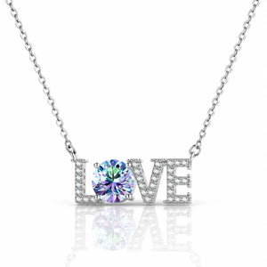 A silver-tone necklace featuring the word 'LOVE' with the letter O replaced by a large round iridescent gemstone, set against a clean white background with a soft reflection below.