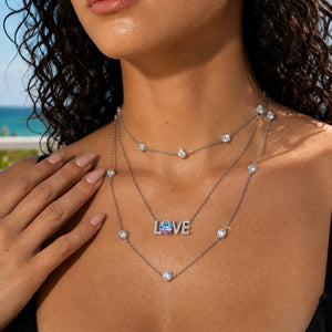 A close-up lifestyle shot of a model wearing a layered silver necklace set. The set includes a short chain with round clear stones, a longer chain with a 'LOVE' pendant featuring a blue heart in the center, and a delicate chain with a single clear stone.