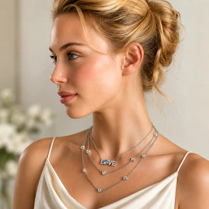 A close-up of a woman in profile wearing a layered silver necklace set. The necklace features a central 'LOVE' initial pendant, small round clear stones, and a delicate chain with tiny silver charms.