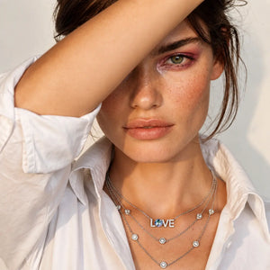 A close-up of a woman in profile wearing a layered silver necklace set. The necklace features a central 'LOVE' initial pendant, small round clear stones, and a delicate chain with tiny silver charms.