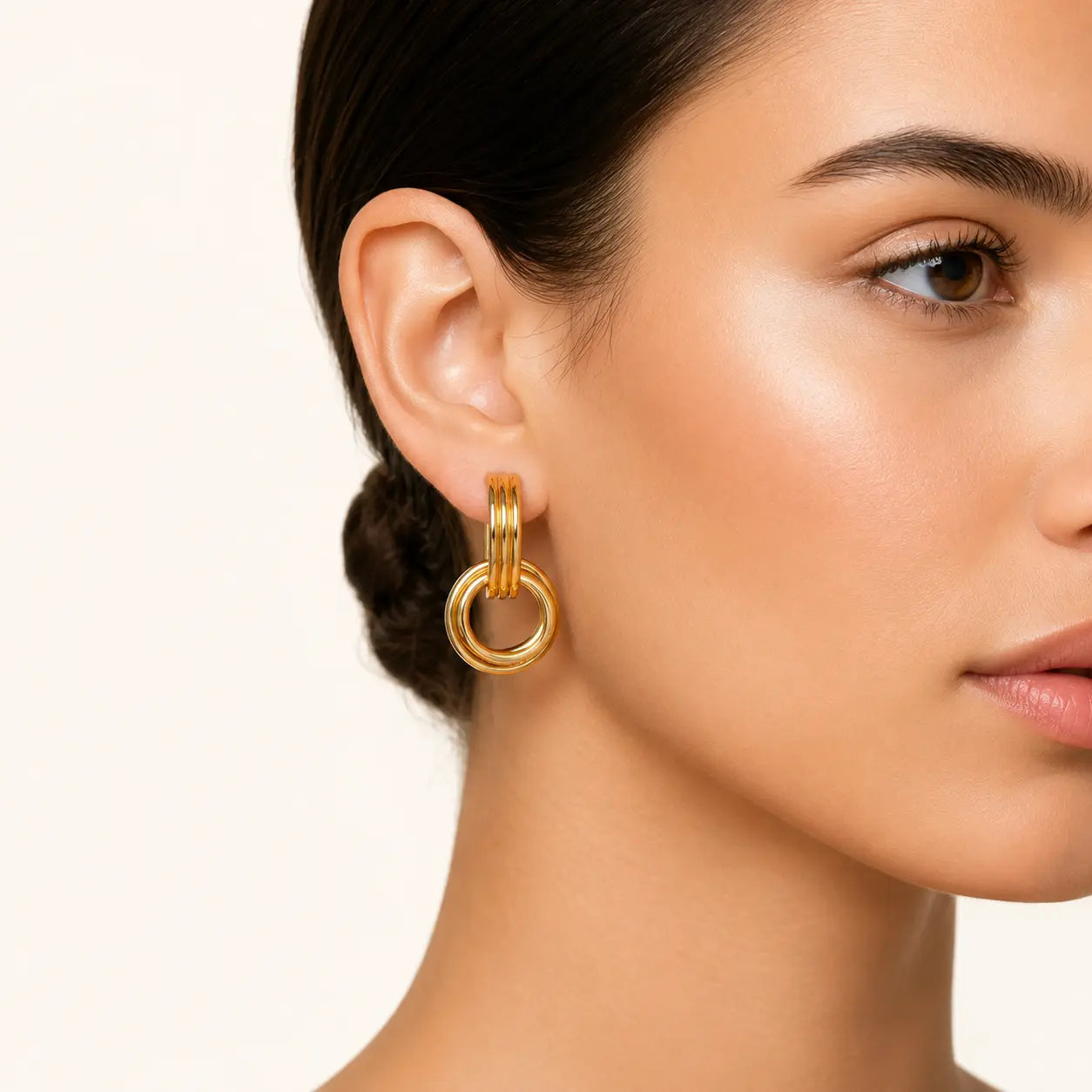 A close-up of a model's ear and side profile wearing a gold huggie hoop earring with a larger interlocking ring drop, against a clean off-white background.